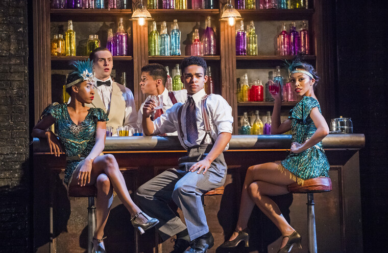 Three people sat on barstools at a bar. Two of them have drinks in hand. A bar tender has a shocked look on his face as a 2nd bar tender leans in to speak to him.
