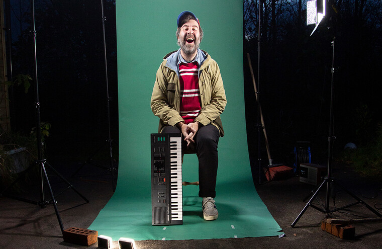 David O'Doherty is sat on a chair outside. A photoshoot set up and camera lights surround him with a green backdrop behind him.