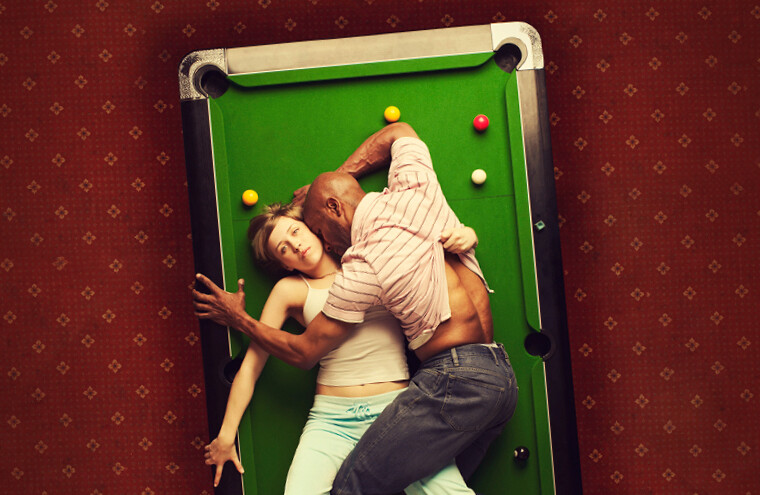 A man lies on top of a woman, laying across a snooker/pool table, as she gazes forward.