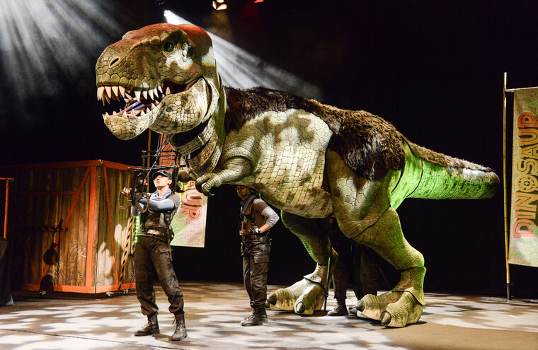 Two people on stage control a large puppet of a T-Rex