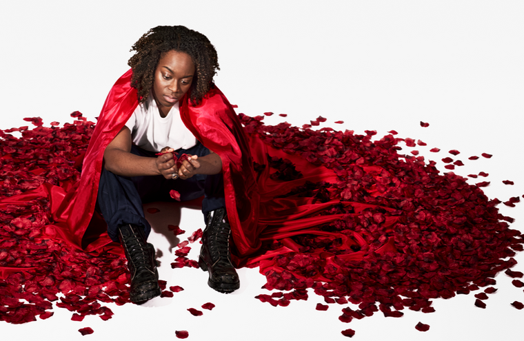 An actor in a red cape sits on the floor surrounded by red petals