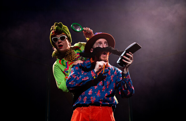 A man with a giant moustache holds a notepad on stage. A woman in sunglasses peers over his shoulder holding a magnifying glass.