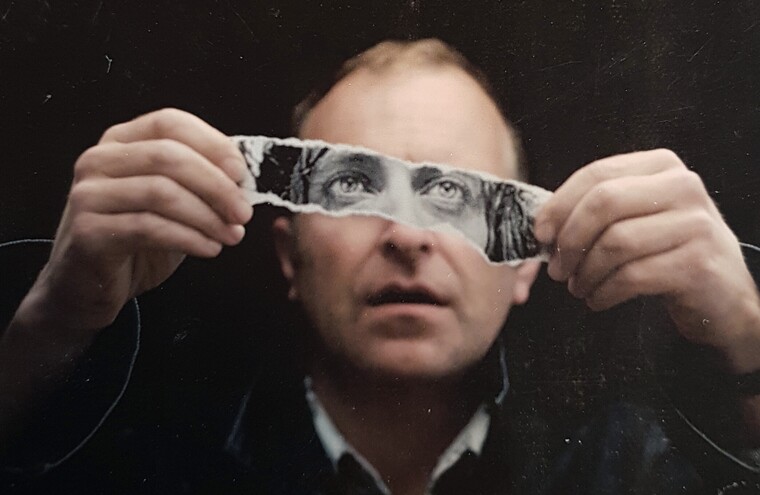 Headshot of Simon Evans - holding up a slip of paper which has a photo of his eyes printed out on, infront of his face.