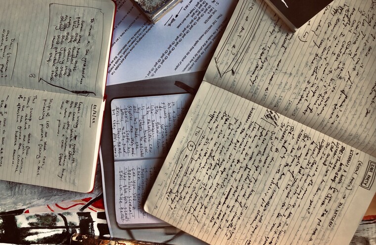 A series of open notebooks laid out on a surface. They have been written in with a black ink pen, books surround them.