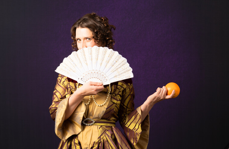 A woman in a Stuart dress holds an open fan over her face and an orange in the other hand.