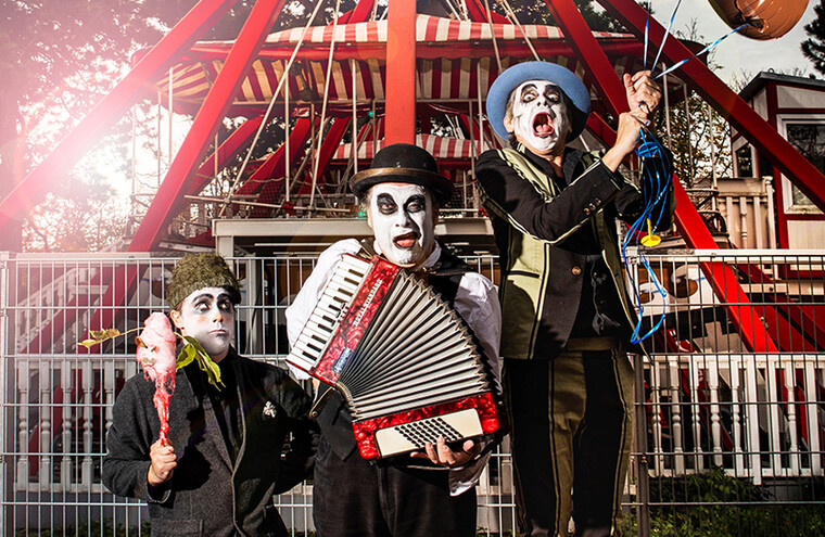 The Tiger Lillies stood in a fairground in front of a ferris wheel. They are dressed in mime makeup. One member is playing the accordion and another is holding a balloon.