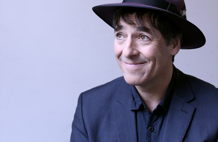 Mark Steel sits on a light grey background in a black suit and purple fedora