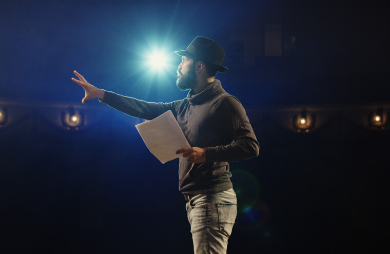 A performer stood on stage with a light shining on him in the background. He is holding a script in one hand with his other arm stretched out in front of him.